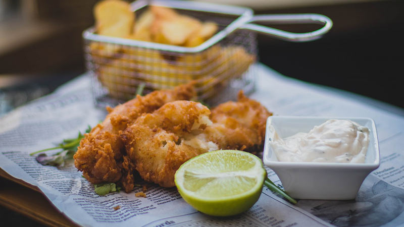 Pollo frito sobre un papel de periódico junto a una cesta con patatas, una salsa blanca y media lima